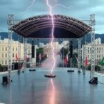 Estrutura de ato político em BH atingida por raio, causando pânico, com céu tempestuoso e montanhas ao fundo.