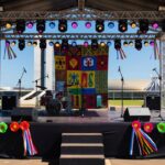 Palco preparado para festa de axé no DF, com equipamentos de som e decorações coloridas, arquitetura de Brasília ao fundo.