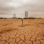 Praça em Brasília com local vazio para ponto de hidratação, arquitetura modernista ao fundo, destacando atraso e riscos à saúde.