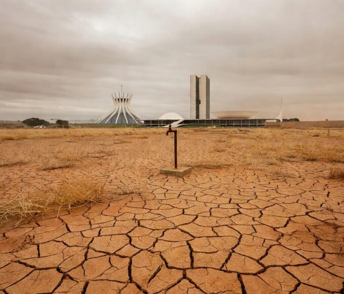 Praça em Brasília com local vazio para ponto de hidratação, arquitetura modernista ao fundo, destacando atraso e riscos à saúde.